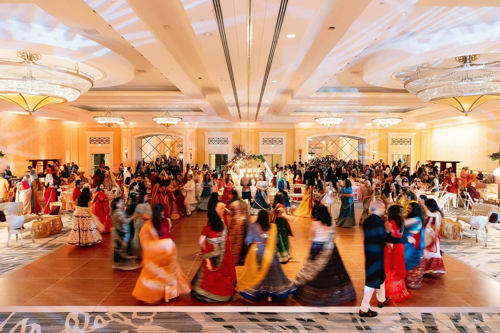 A large group of people in formal attire dancing at an event venue with chandeliers and decorative lighting.